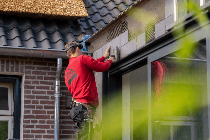 Werkman plaatst grijze tegels op een buitenmuur naast een raam tijdens renovatiewerk aan een woning.