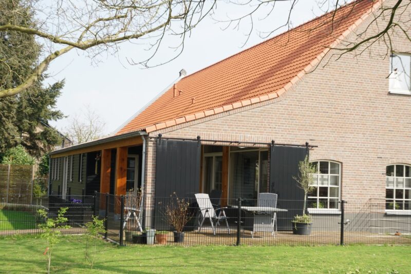 Detail van de aanbouw van een gerenoveerde woning in Cuijk, met robuuste houten staanders, zwarte gepotdekselde planken en grote zwarte schuifdeuren bij het overdekte terras, gerealiseerd door Claassen Bouw.