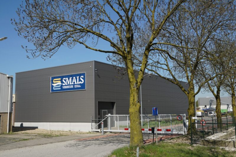 Vooraanzicht van het moderne bedrijfspand van Smals Verhuur in Katwijk, gebouwd door Claassen Bouw, voorzien van donkergrijze geribbelde gevelbeplating en een beveiligde toegangspoort.