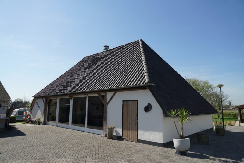 Zijaanzicht van een vernieuwd campinggebouw in Beers, verbouwd door Claassen Bouw, met een strak wit gestucte gevel, houten details, grote glazen deuren en een pannendak op een zonnige dag.
