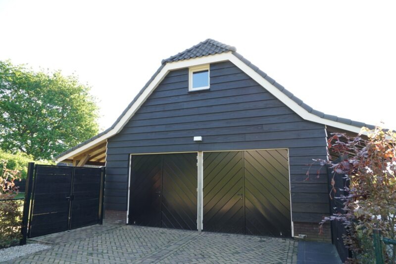 Close-up van de voorzijde van een houten bijgebouw gebouwd door Claassen Bouw, met strakke donkere gevelplanken en brede dubbele garagedeuren met een diagonaal plankmotief.