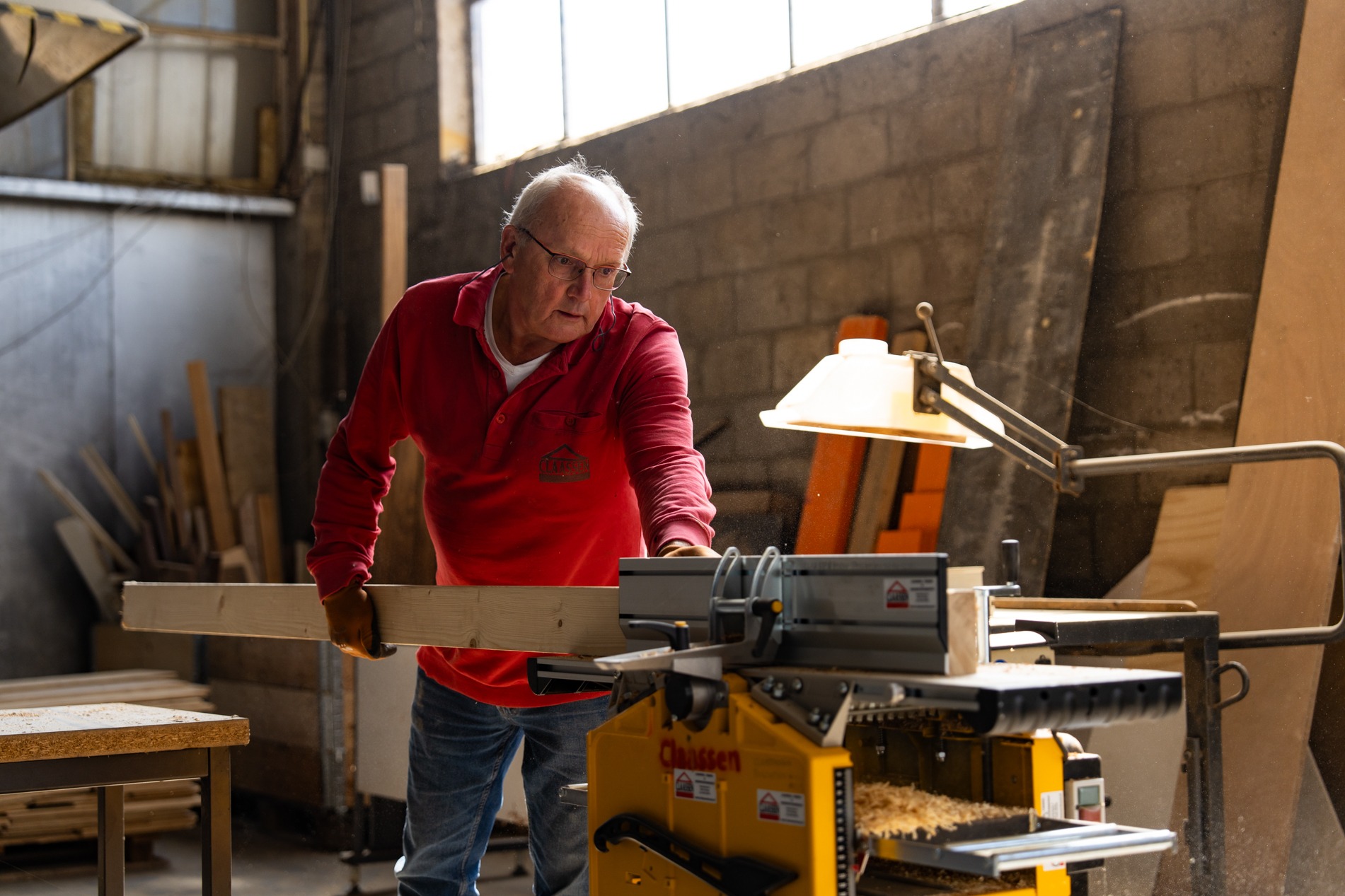 Een ervaren vakman van Claassen Bouw in een rode polo bewerkt een lange houten balk met een vlakbank in een werkplaats, verlicht door een gerichte werklamp.