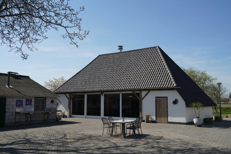 Vooraanzicht van een gerenoveerd wit campinggebouw in Beers door Claassen Bouw, gekenmerkt door een grote glazen pui omlijst met houten spanten, een donker pannendak en een klein terras.
