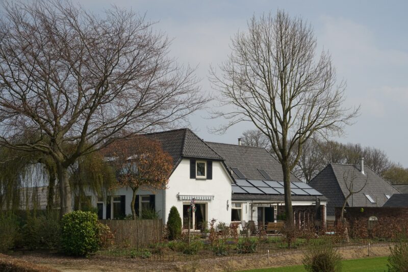 Gerestaureerd wit woonhuis bij een camping in Beers, met donkere luiken, zonnepanelen op het dak en een weelderige voortuin met grote bomen.