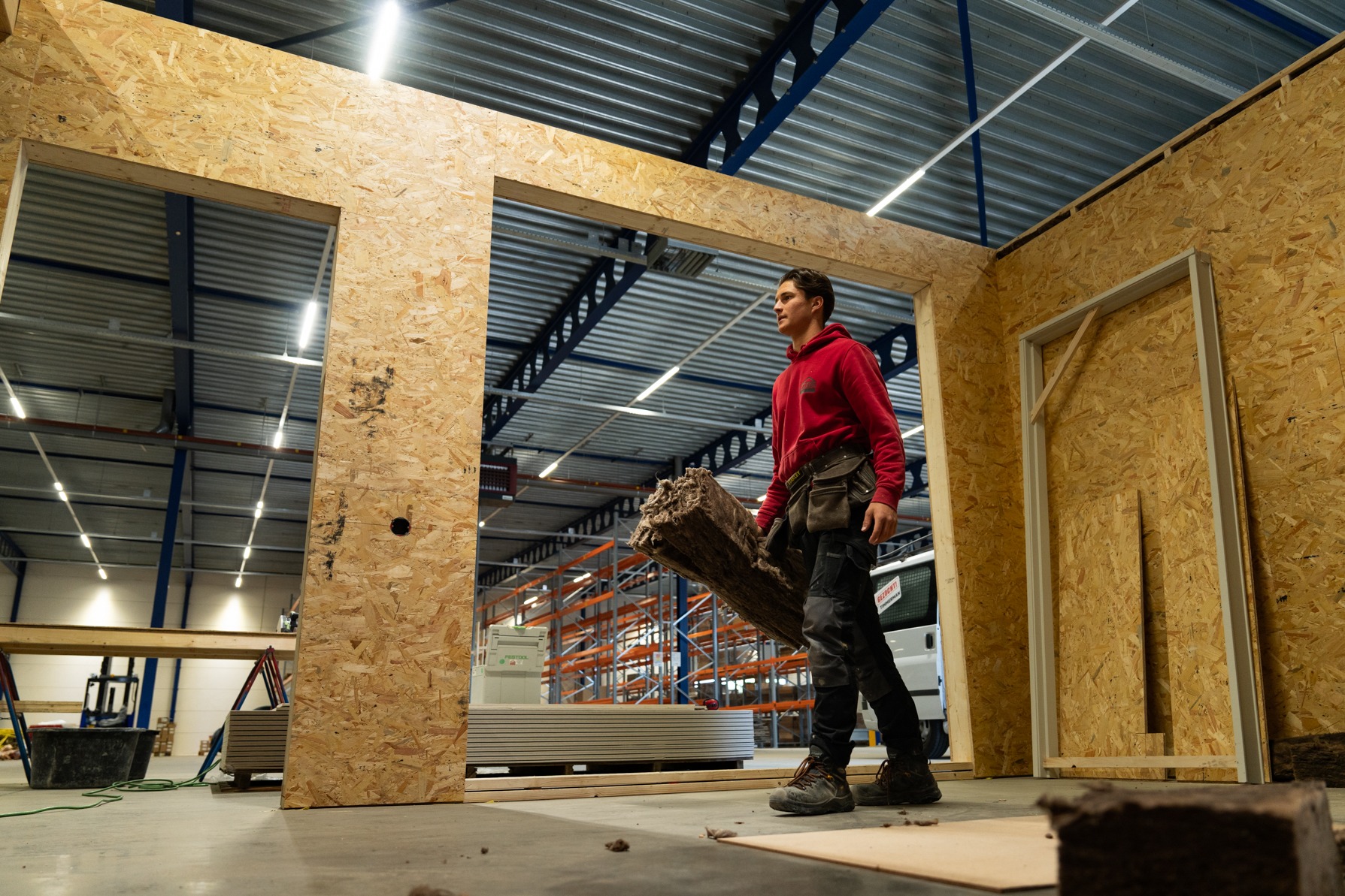Een jonge medewerker van Claassen Bouw in rode werkkleding draagt een bundel isolatiemateriaal in een prefabricage-werkplaats, omringd door houten HSB-wandframes gemaakt van OSB-platen en achterliggende palletstellingen.
