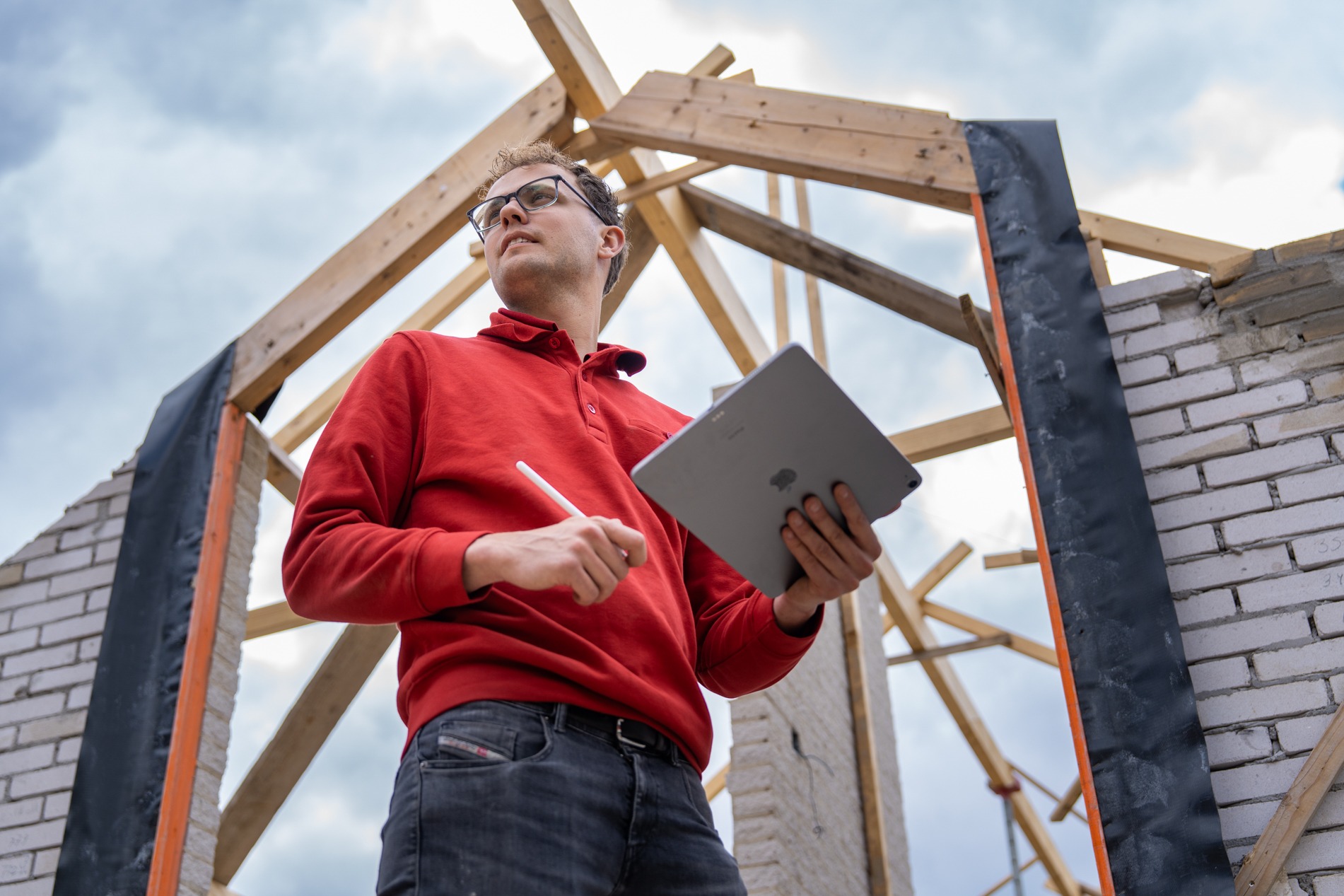 Wouter Nijhuis van Claassen Bouw inspecteert een woning in aanbouw met een tablet en stylus in zijn hand, staand tussen gemetselde muren onder een houten kapconstructie.