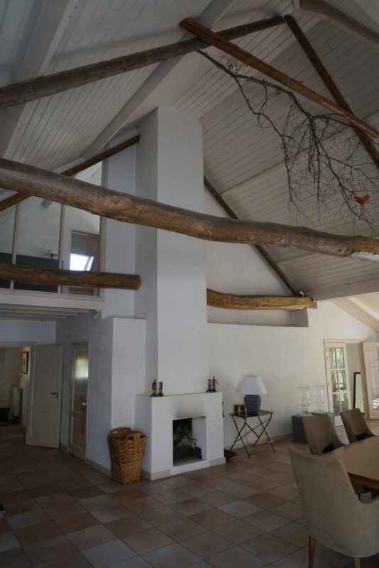 Interieur van een landelijke woning gerenoveerd door Claassen Bouw, met een strakke witte schouw, indrukwekkende authentieke houten draagbalken over de hele breedte en een lichte tegelvloer.