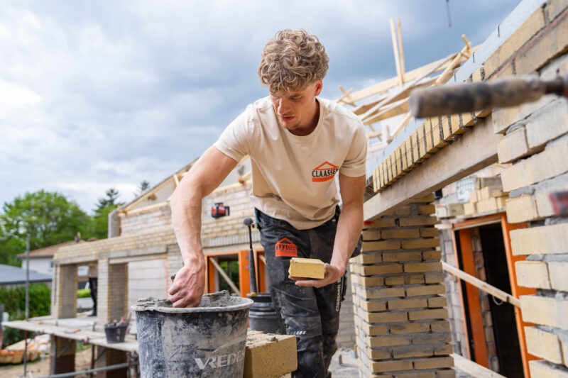 Een jonge metselaar van Claassen Bouw mengt cement in een zwarte emmer met zijn linkerhand terwijl hij een enkele baksteen in zijn rechterhand vasthoudt op een bouwplaats.