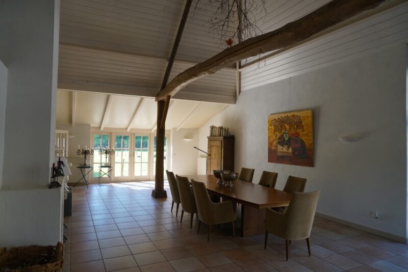 Lichte leefruimte in een verbouwde boerderij van Claassen Bouw, gekenmerkt door een hoge nok, een verticale houten staander, een grote eettafel en veel natuurlijk licht door de openslaande tuindeuren.