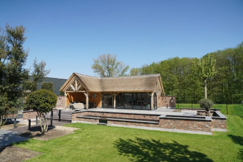 Vrijstaand eikenhouten poolhouse in Boxmeer met een rieten kap, een open overkapping met loungehoek en grote glazen puien, gebouwd op een verhoogd terras aan een strak afgewerkt zwembad met gemetselde randen.