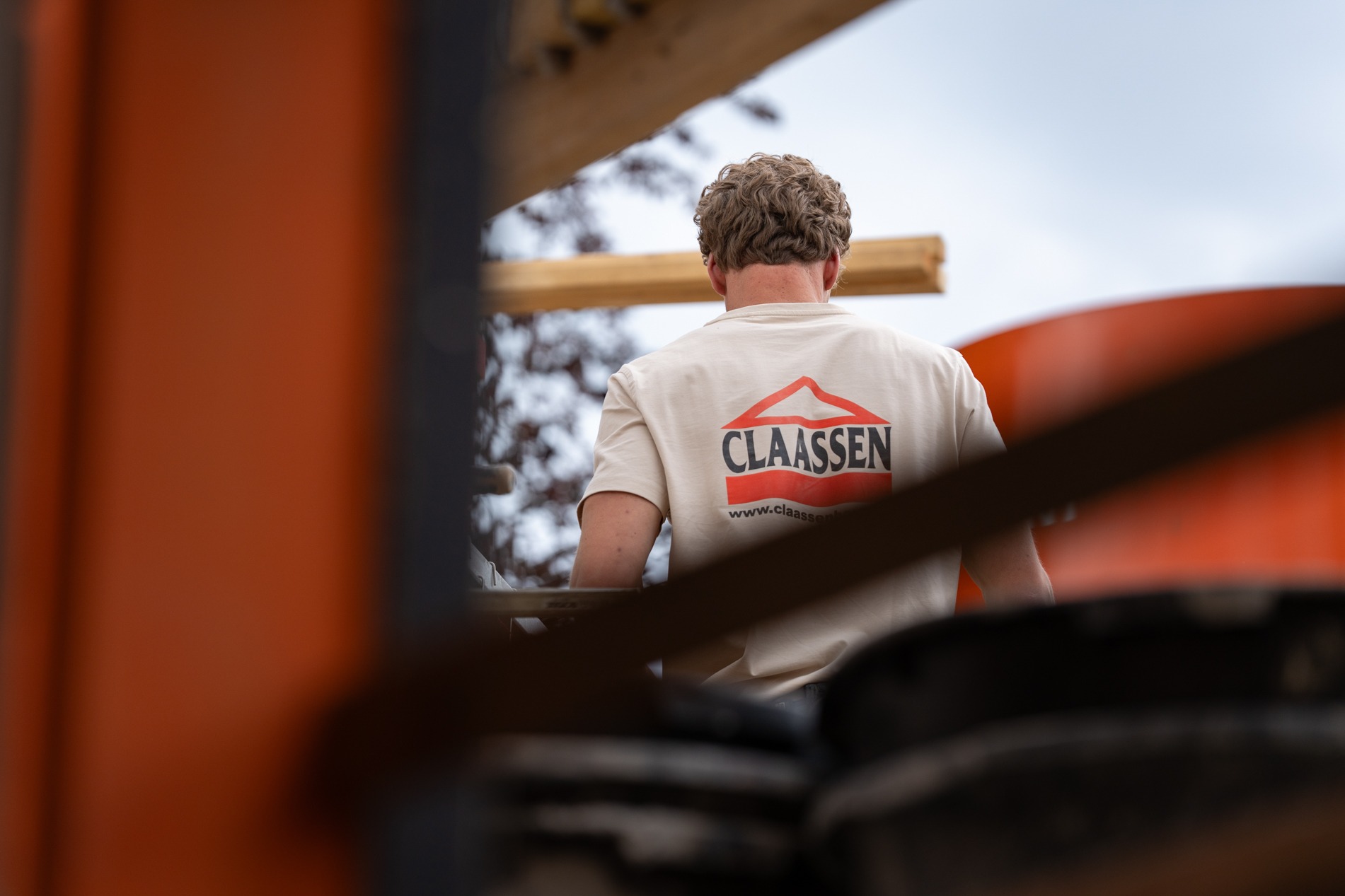 Achteraanzicht van een bouwvakker op de bouwplaats, waarbij het opvallende rode bedrijfslogo van Claassen Bouw prominent zichtbaar is op de rug van zijn beige werkshirt.