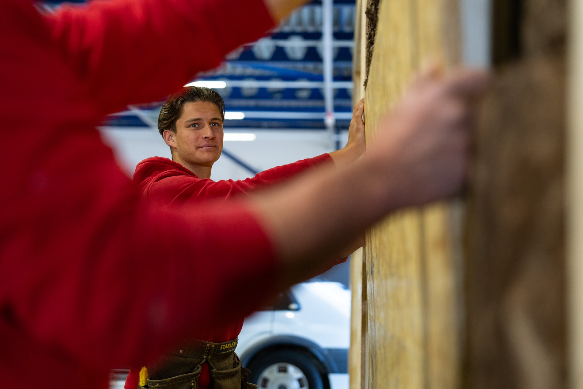 Een jonge medewerker van Claassen Bouw in een rode trui werkt samen met een collega, zichtbaar in de voorgrond, om een grote houten wandplaat te bevestigen in een werkplaats.