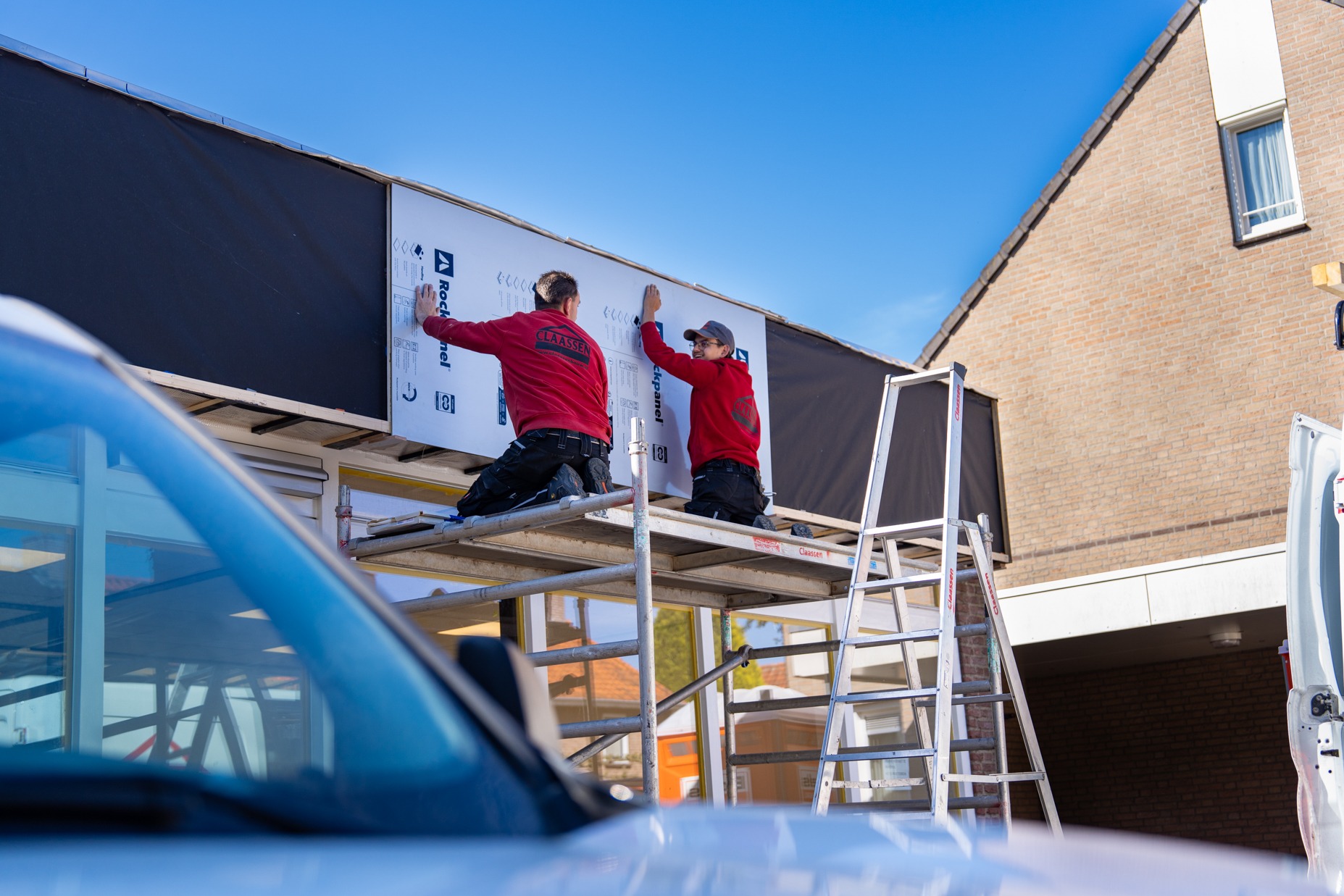 Twee medewerkers van Claassen Bouw in rode werkkleding installeren vanaf een steiger witte Rockpanel gevelbekleding aan de buitenkant van een bedrijfspand.