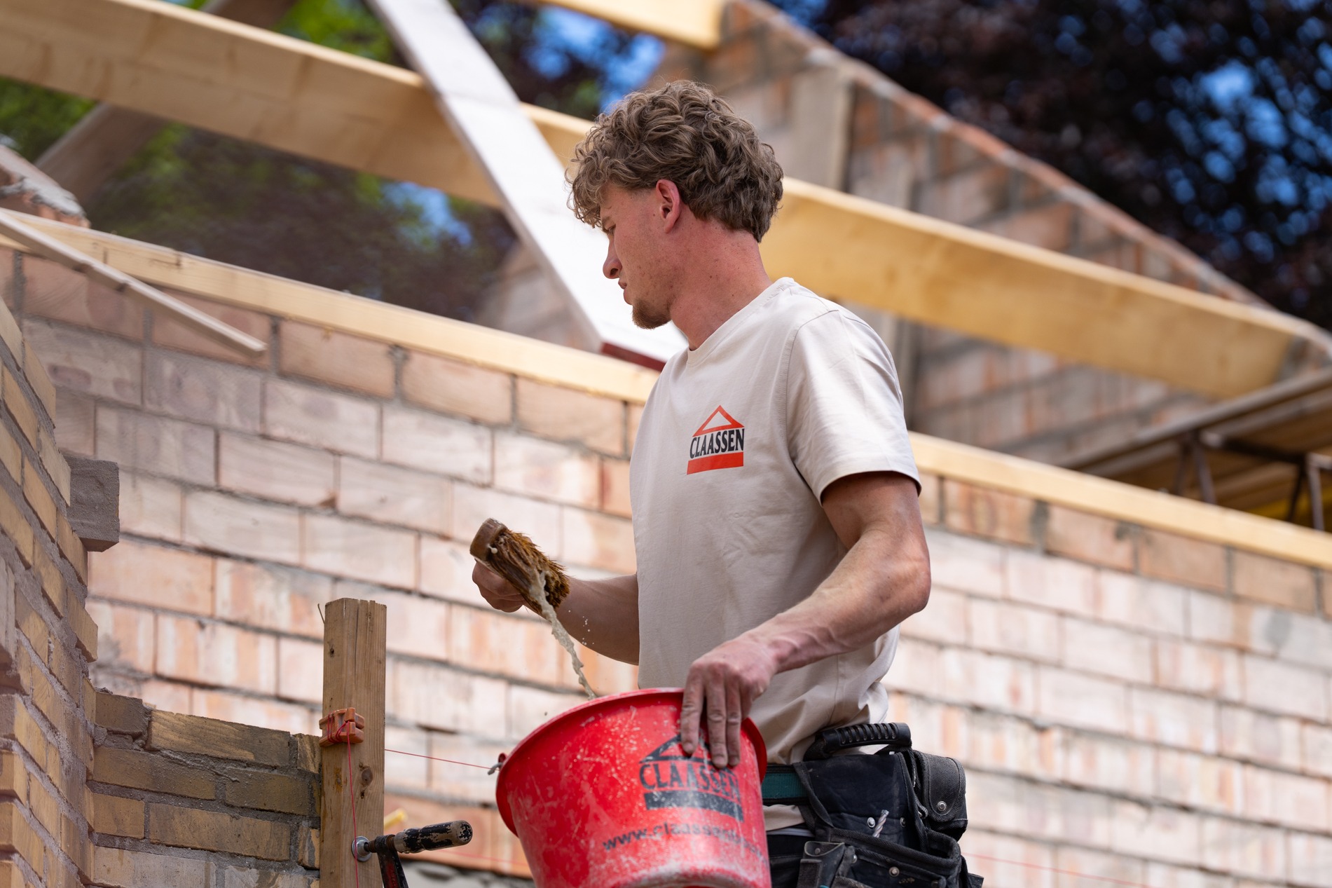 Een jonge bouwvakker van Claassen Bouw bevochtigt een bakstenen muur met een blokkwast vanuit een rode emmer tijdens werkzaamheden op de bouwplaats.