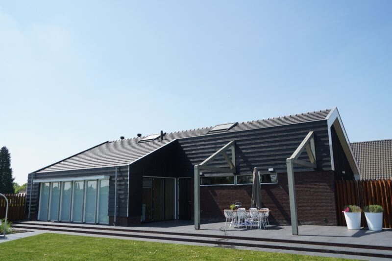 Tuinzijde van een modern gebouw door Claassen Bouw, met deels zwarte houten en deels bakstenen gevels, grote glazen schuifdeuren en een betegeld terras met een robuuste houten pergola.