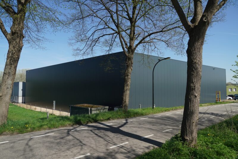Zijaanzicht van een strakke, moderne bedrijfshal in Katwijk gerealiseerd door Claassen Bouw, afgewerkt met grote donkergrijze sandwichpanelen, rustig gelegen achter een rij bomen aan een doorgaande weg.