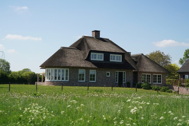 Landelijke bakstenen villa met een golvend rieten dak en klassieke witte kozijnen, gefotografeerd op een zonnige dag vanuit een bloeiend weiland in Beers.