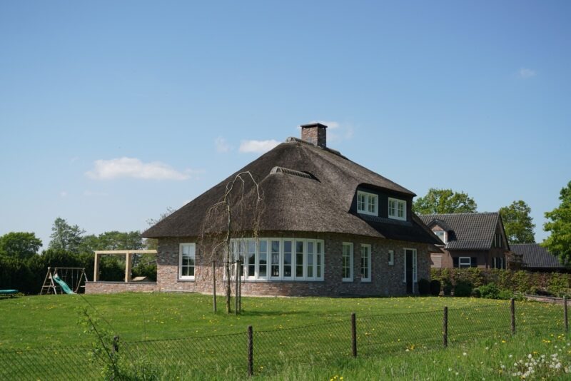 Zijaanzicht van een landelijke nieuwbouwwoning in Beers met een prominente rieten kap, een halfronde serre met witte kozijnen en een houten overkapping in de zijtuin.