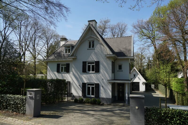 Luxe witte nieuwbouw villa in Nijmegen gerealiseerd door Claassen Bouw, prachtig gelegen tussen de bomen, met klassieke details zoals donkere raamluiken en een bijpassende garage op de achtergrond.