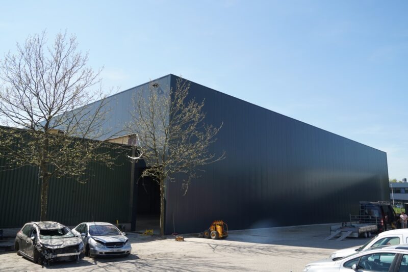 Grote, nieuw gebouwde donkergrijze bedrijfshal van Claassen Bouw in Katwijk, naadloos aangesloten op een bestaand groen pand, met een open toegang en enkele schadeauto's op het voorterrein.
