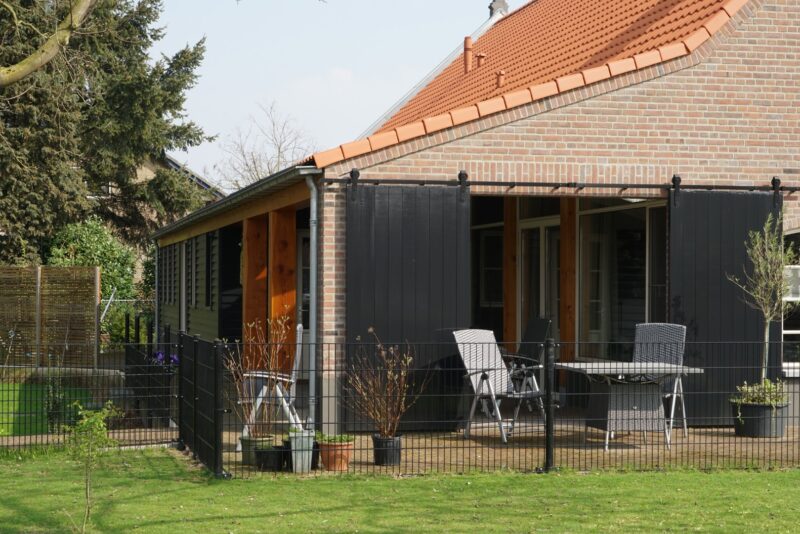 Detailopname van het overdekte buitenterras bij een door Claassen Bouw gerenoveerde woning in Cuijk, met robuuste houten staanders, grote zwarte schuifdeuren op een railsysteem en een gezellige zithoek.