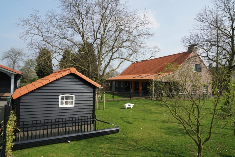 Overzicht van een ruime groene tuin in Cuijk met een door Claassen Bouw gerenoveerde woonboerderij, een lange zwarte houten aanbouw en een bijpassend speel- of tuinhuisje op de voorgrond.