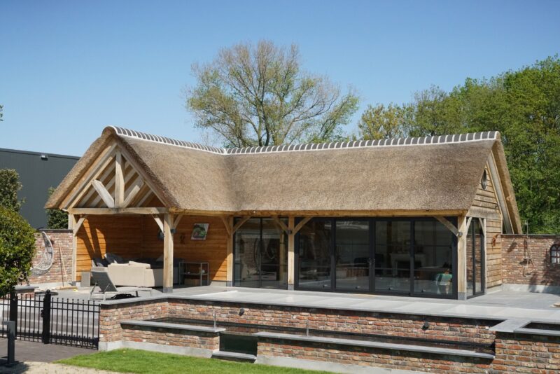 Buitenaanzicht van een luxe eikenhouten poolhouse in Boxmeer, voorzien van een ambachtelijke rieten kap, een open loungeterras en strakke zwarte glazen schuifpuien, grenzend aan het zwembad.