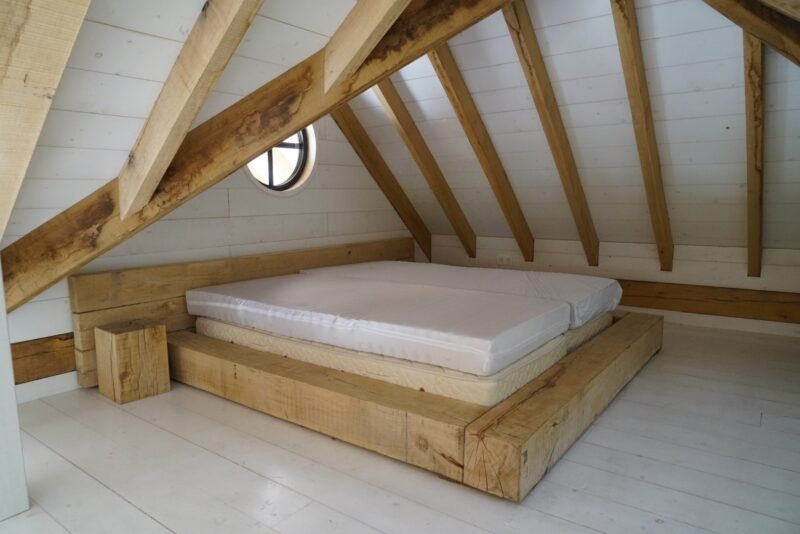 Slaapvide in een door Claassen Bouw gerealiseerd poolhouse in Boxmeer, met een op maat gemaakt robuust houten bedframe onder een schuin dak met zichtbare eikenhouten spanten en witte wanden.