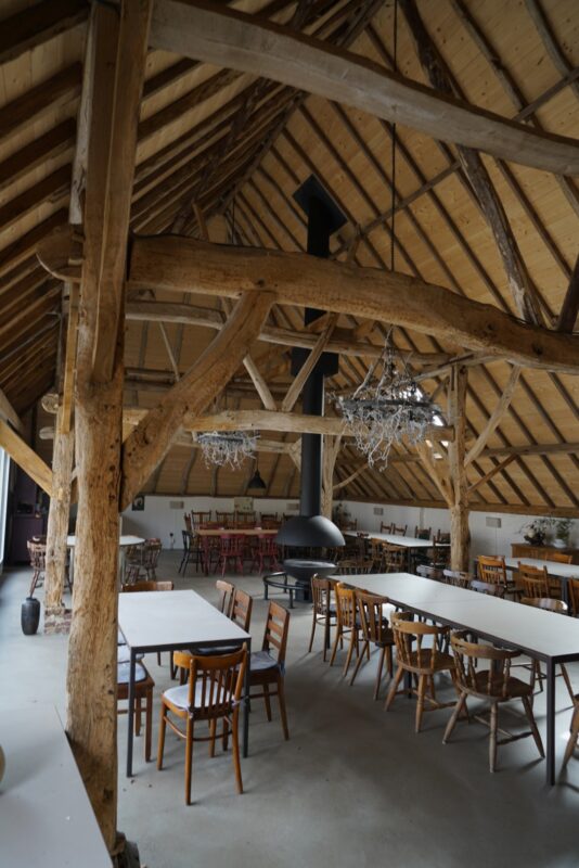 Sfeervolle recreatieruimte op een camping in Beers met robuuste houten gebinten, tafels met stoelen en een opvallende centrale hangende open haard, gerenoveerd door Claassen Bouw.