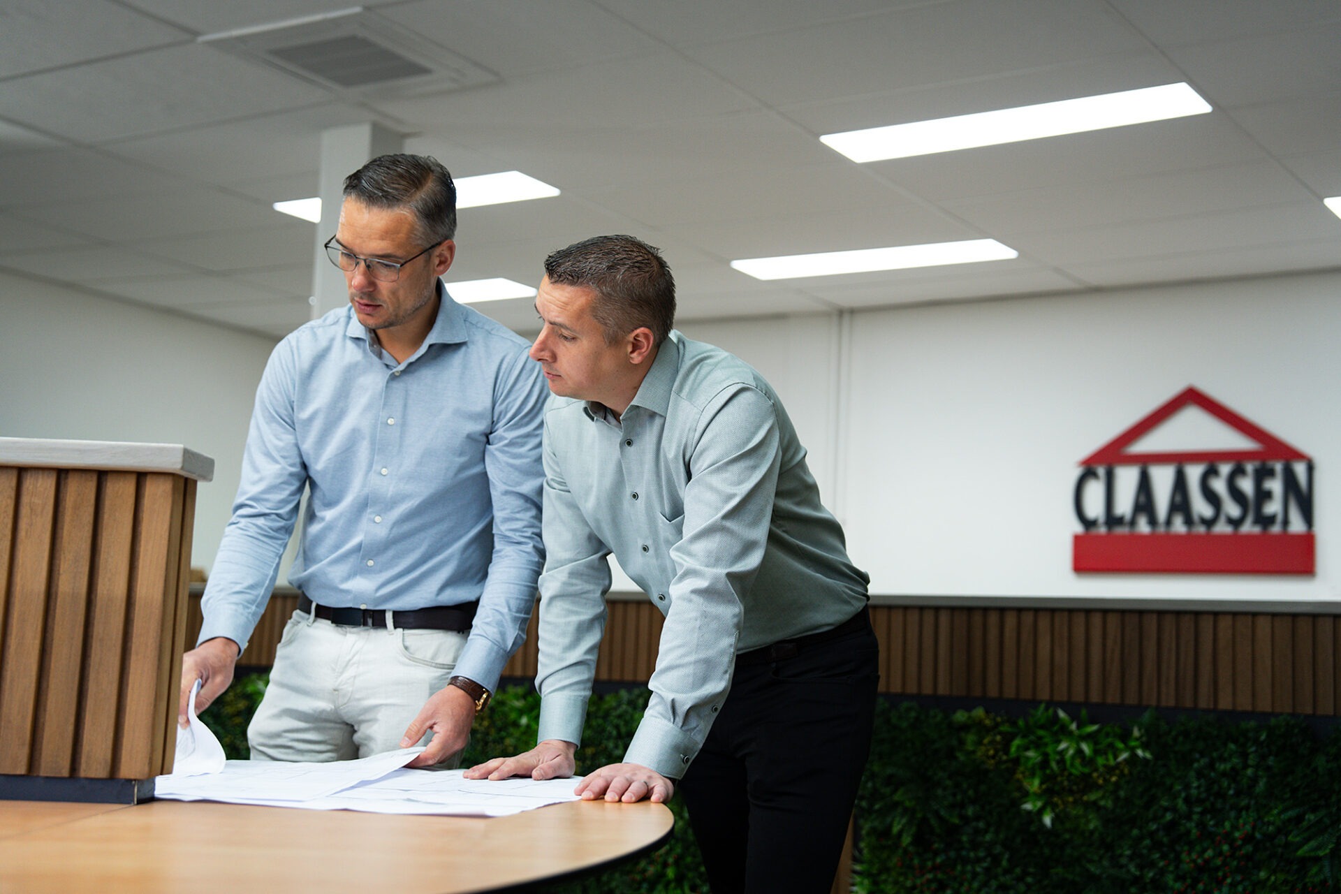René en Paul Claassen bestuderen samen aandachtig papieren bouwtekeningen aan een tafel in het kantoor van Claassen Bouw, met het bedrijfslogo zichtbaar op de achtergrond.