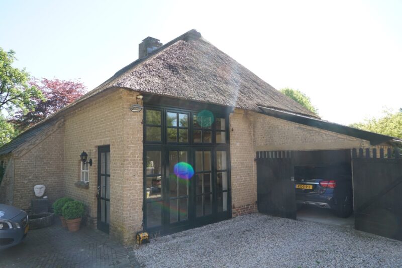 Gerenoveerd stenen bijgebouw met rieten kap door Claassen Bouw, getransformeerd met grote zwarte pui-deuren van glas en een openstaande inpandige garage aan een grindoprit.