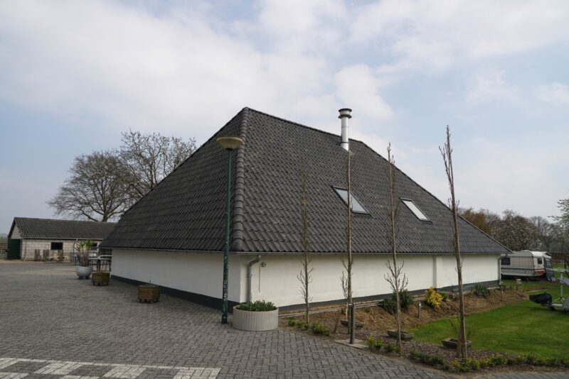 Achteraanzicht van een ruim, wit stenen gebouw met een fors donker pannendak en dakramen op een camping in Beers, gerenoveerd door Claassen Bouw.