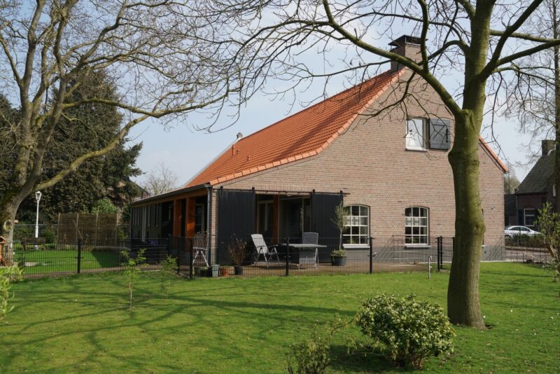 Achterzijde van een gerenoveerde woonboerderij in Cuijk door Claassen Bouw, met een combinatie van traditioneel metselwerk en zwart hout, een opvallend rood pannendak en een overdekt terras met zwarte staldeuren.