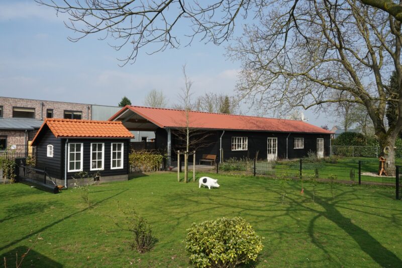 Gerenoveerd langwerpig zwart houten bijgebouw met een rood pannendak en een bijpassend tuinhuisje in een ruime tuin met gazon, verbouwd door Claassen Bouw in Cuijk.
