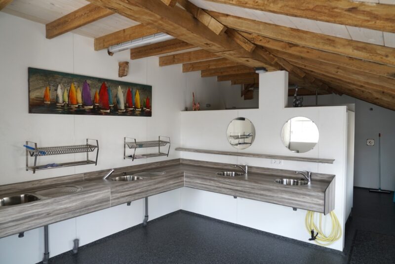 Modern en strak afgewerkte wasruimte met zichtbaar houten balkenplafond op een camping in Beers, voorzien van rvs-wasbakken verwerkt in een houtlook blad, gerealiseerd door Claassen Bouw.