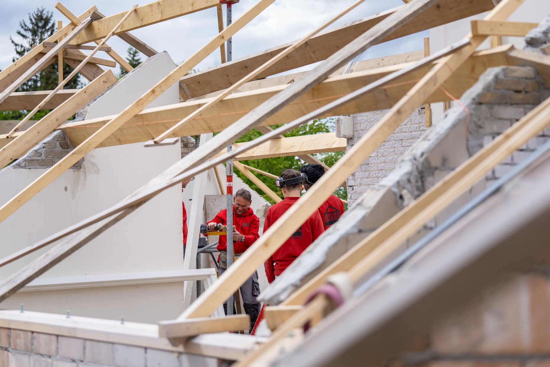 Een team van bouwvakkers van Claassen Bouw in rode werkkleding werkt samen aan de houten kapconstructie van een woning in aanbouw, waarbij één vakman een accuboor gebruikt.