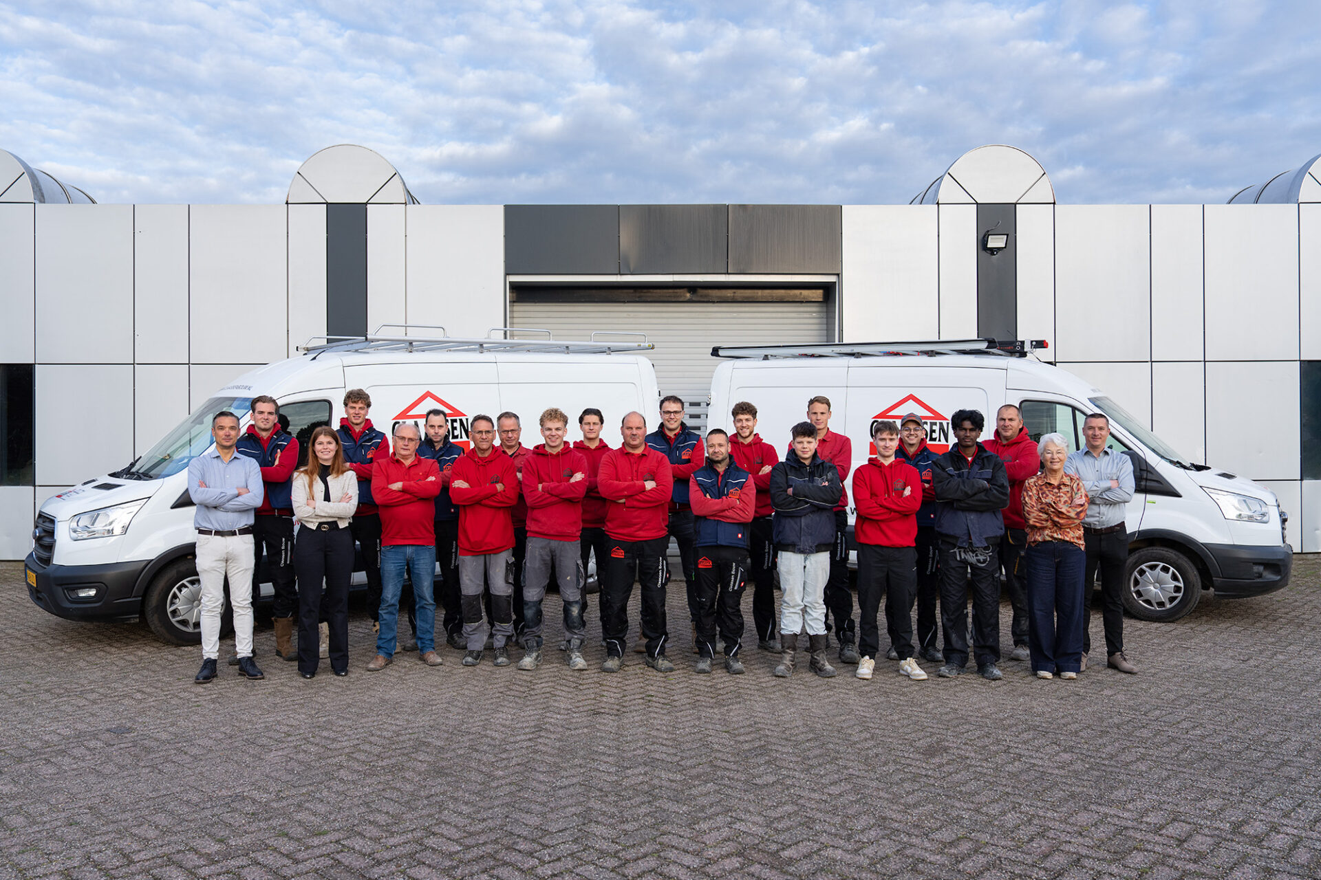Groepsfoto van het voltallige personeel van Claassen Bouw, met de familie en medewerkers trots poserend voor het bedrijfspand en twee witte bedrijfswagens.