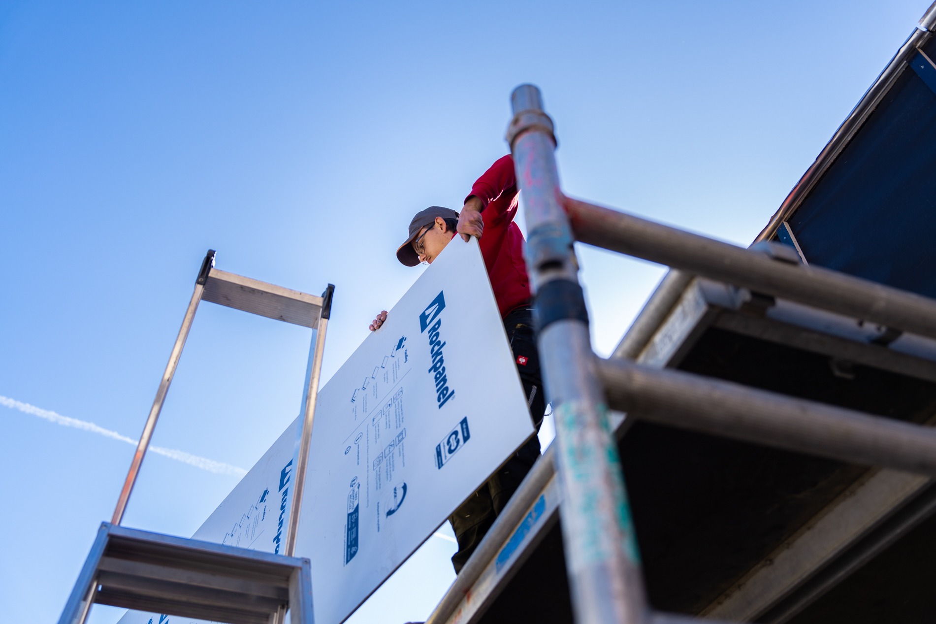 Laag standpunt van een vakman van Claassen Bouw op een steiger, die een grote witte Rockpanel plaat vasthoudt voor montage tegen een strakblauwe lucht.