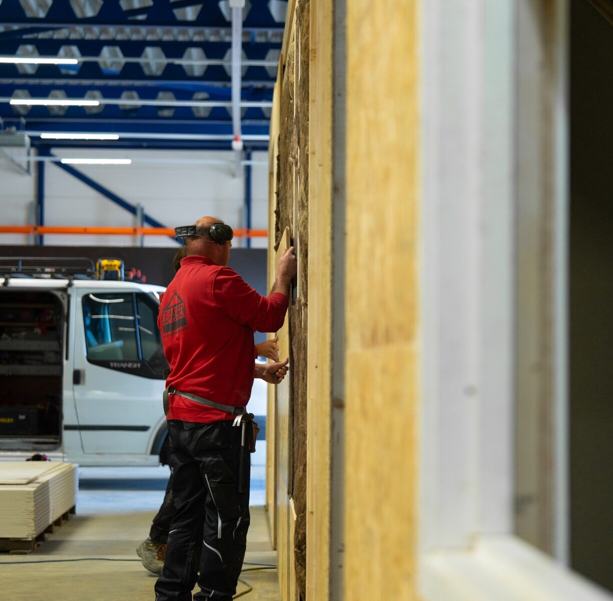 Een vakman van Claassen Bouw in een rode polo en met gehoorbescherming plaatst houten panelen tegen een geïsoleerde wand in de werkplaats, terwijl een collega op de voorgrond zijn telefoon bekijkt.