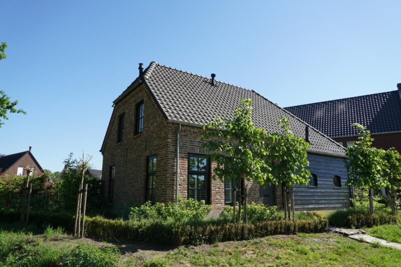 Vooraanzicht van het gemetselde gedeelte van een woning gebouwd door Claassen Bouw, gekenmerkt door landelijke bakstenen, donkere raamkozijnen en jonge leibomen in de voortuin.