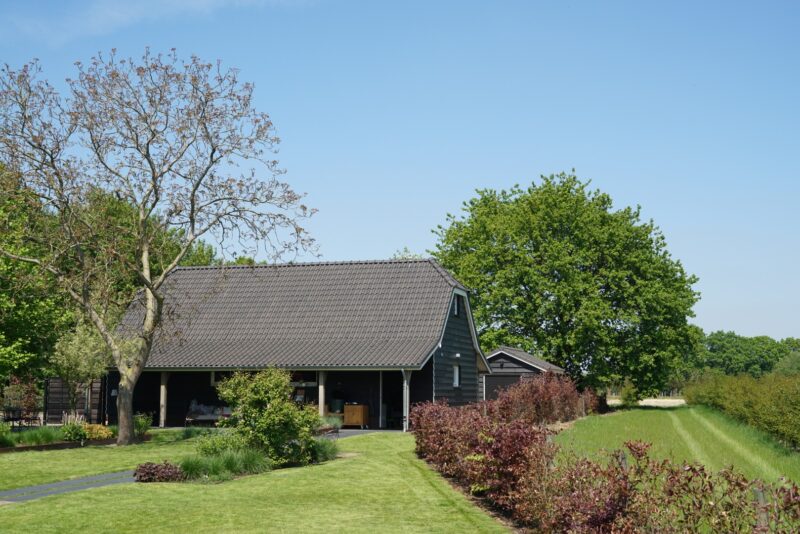 Vrijstaand houten bijgebouw in een weelderige groene tuin, gebouwd door Claassen Bouw, voorzien van donkere gevelbekleding, een pannendak en een ruime overdekte veranda.