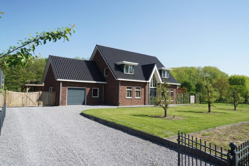 Moderne vrijstaande nieuwbouwwoning van Claassen Bouw met donkere bakstenen, een donker pannendak, een opvallende entree met een hoge glaspartij, een inpandige garage en een ruime oprit van grind.