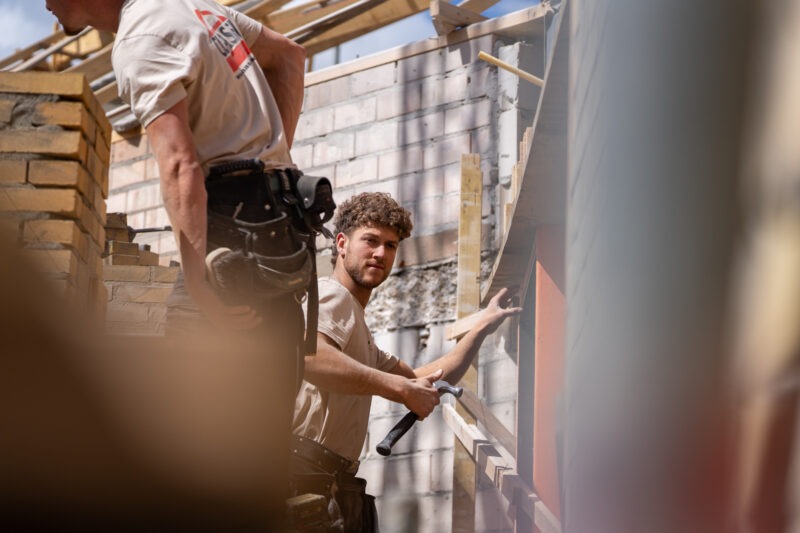 Twee bouwvakkers van Claassen Bouw op een bouwplaats; de voorste draagt een volle gereedschapsriem, de achterste kijkt naar hem en houdt een hamer en breekijzer vast bij een houten frame.