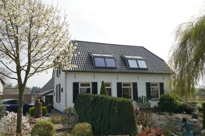 Vooraanzicht van een wit geschilderd landhuis in Beers met dakkapellen, donkere raamluiken en een sfeervolle tuin, gefotografeerd naast een bloeiende magnoliaboom.