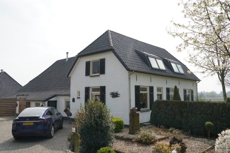 Karakteristiek wit woonhuis met donkere luiken en strakke dakkapellen bij een camping in Beers, verbouwd door Claassen Bouw, met een oprit waar een blauwe Tesla geparkeerd staat.