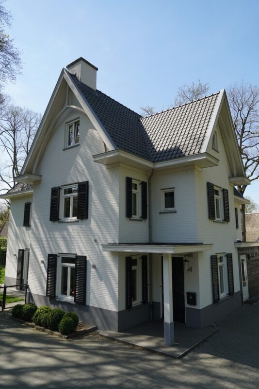 Zijaanzicht van een riante witte nieuwbouwwoning in Molenhoek gebouwd door Claassen Bouw, met focus op de overdekte entree, kruiskozijnen met donkere luiken, dakoverstekken en de opvallende grijze gevelplint.