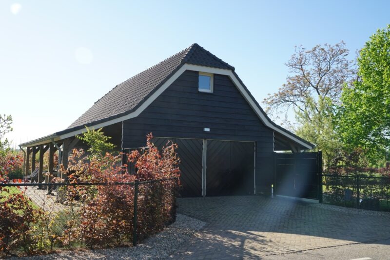Aanzicht van een zwart houten bijgebouw van Claassen Bouw met een donker pannendak, grote dubbele garagedeuren, een overkapping aan de zijkant en een bestrate oprit.